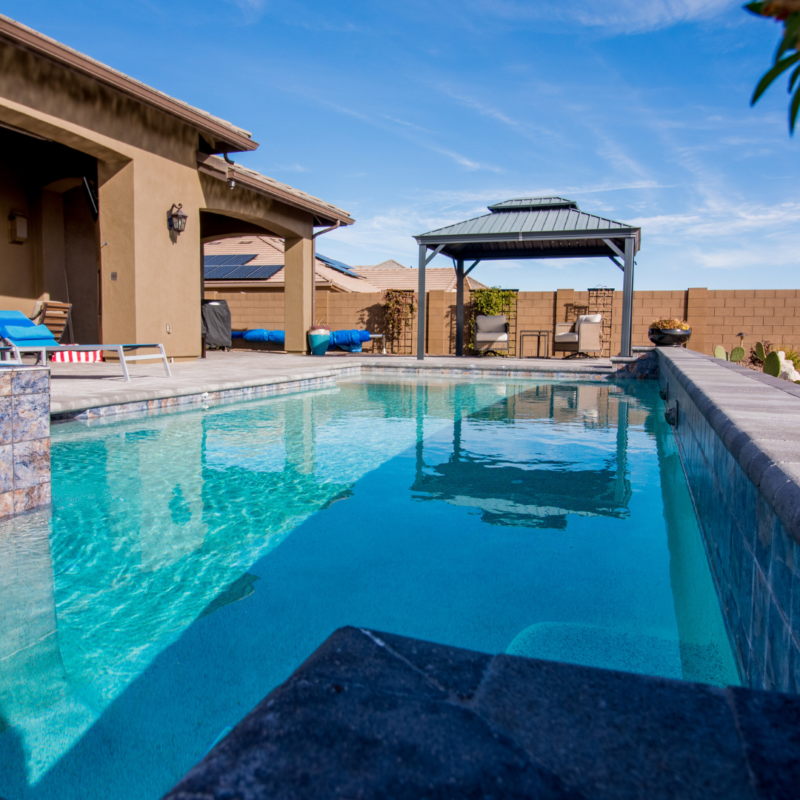 A picturesque backyard scene at the Hutchinson residence features a clear swimming pool surrounded by a stone deck, lounge chairs, and a covered gazebo. A wooden fence and the house are visible in the background under a clear blue sky.