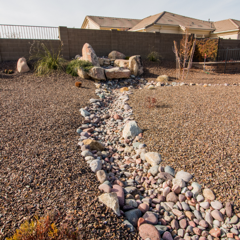 The Hutchinson Residence features a landscaped backyard with a dry creek bed made of various sized pebbles and rocks winding through a gravel-covered area. Sparse plants and rocks are scattered around, and a brown brick wall with metal fencing surrounds the yard. Several houses are visible in the background.