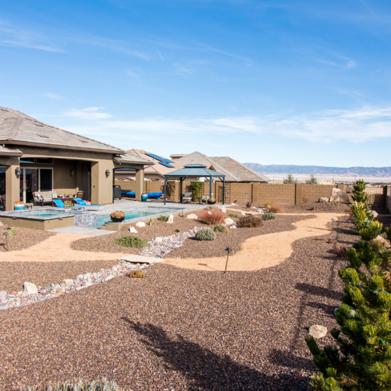 The Hutchinson Residence features a modern backyard with a covered patio, swimming pool, and shaded seating area. The yard is landscaped with gravel, rocks, and drought-resistant plants. A fence and mountains are visible in the background under a clear blue sky.