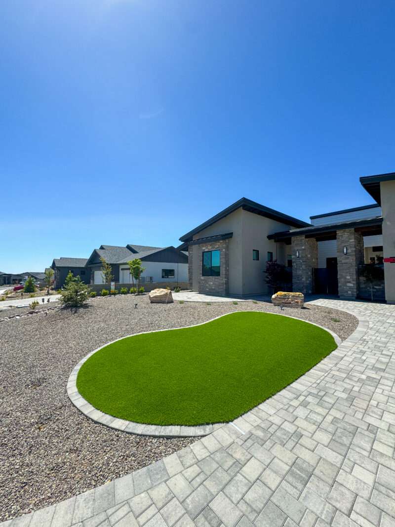 The Wong Residence is a modern single-story house with a stone facade under a clear blue sky. The front yard features a kidney-shaped patch of artificial grass surrounded by gravel and two small rocks, with a paved walkway leading to the entrance. Nearby houses are visible in the background.