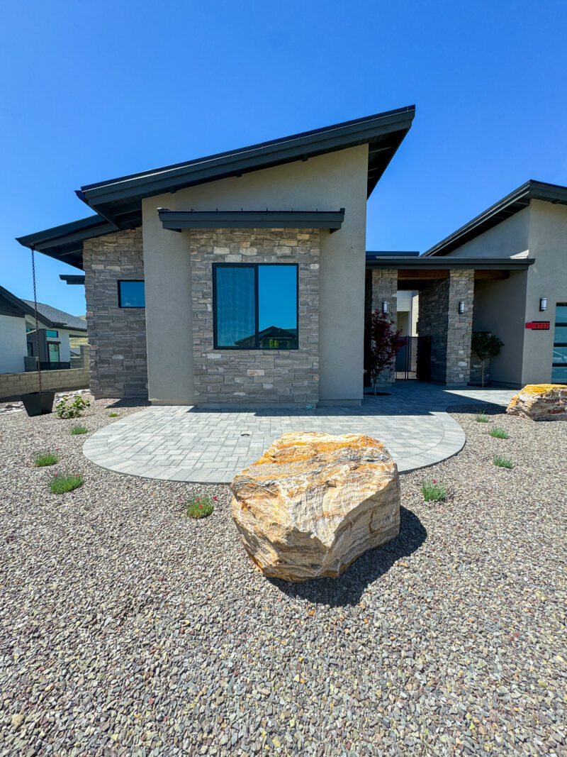 The Wong Residence is a modern house with a light gray stone exterior and a dark roof, featuring a round paved area in front. The landscape includes gravel and a large decorative rock in the foreground, with clear blue skies overhead.