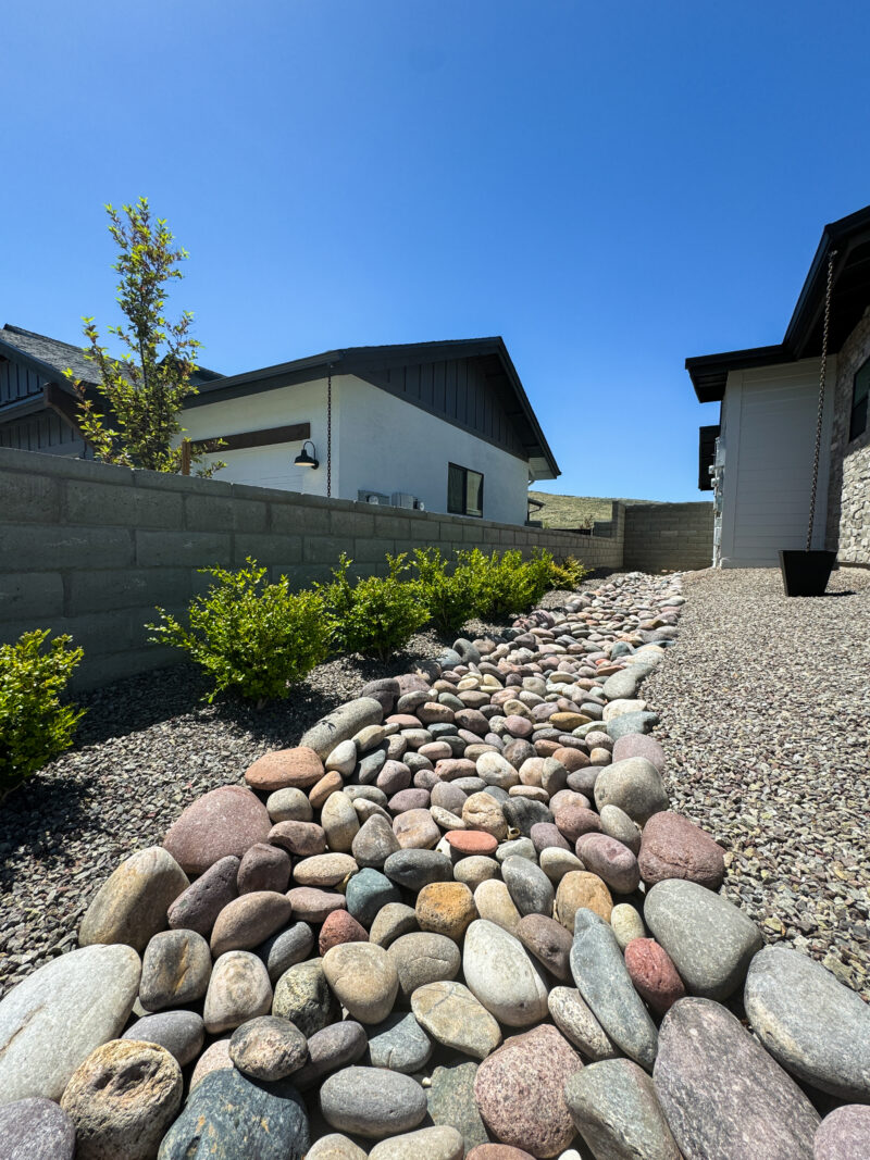 A backyard landscaped at the Wong Residence features a dry rock bed with various sizes and colors of smooth stones. Low greenery lines the rock bed, and in the background, houses with modern architectural designs sit under a clear blue sky.