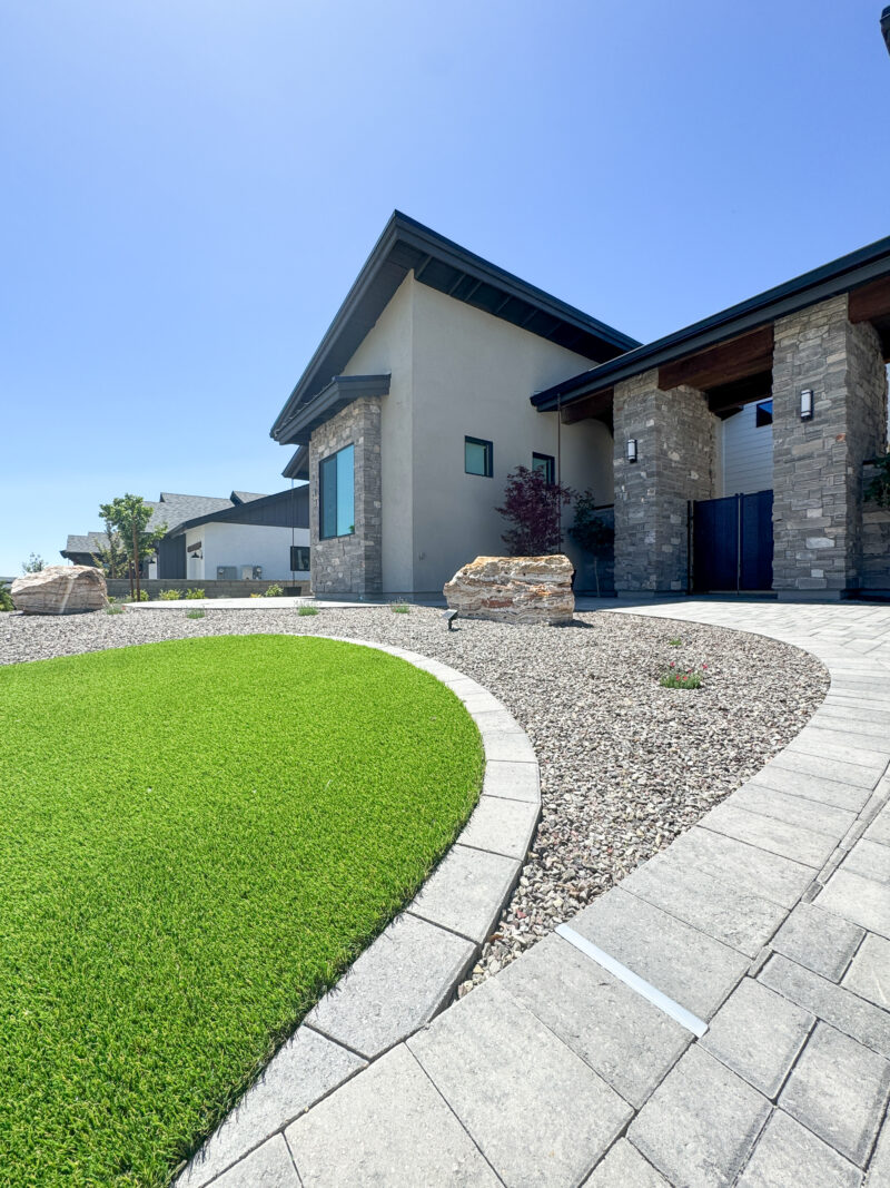 The Wong Residence is a modern house with a mix of stone and stucco exterior, featuring a well-maintained front yard. The yard boasts green artificial grass, decorative rocks, and a curved paved walkway leading to the front entrance under the clear blue sky.