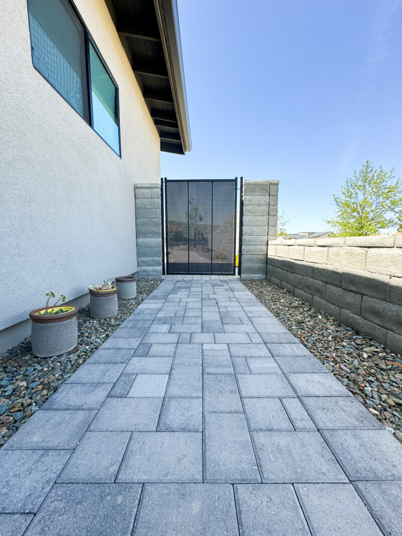 A paved walkway leading to a black metal gate with stone pillars is flanked by a beige house on one side and a brick wall on the other. There are three small planters with plants on the left, and beyond the gate, a tree is visible against a clear blue sky at the Wong Residence.