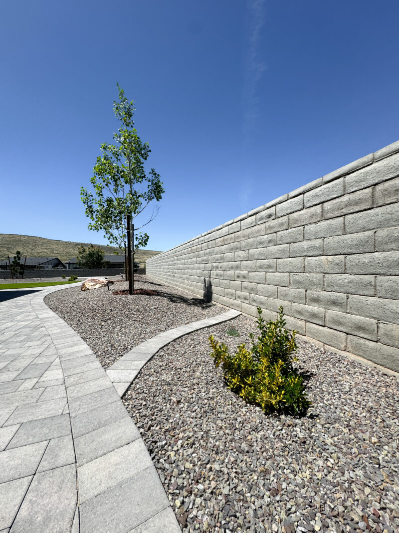 The landscaped area at the Wong Residence features a young tree and a small shrub planted in a bed of rocks. A light-colored brick retaining wall lines the right side, while a paved walkway curves through the area. The sky is clear and blue, adding to the serene ambiance of the residence.