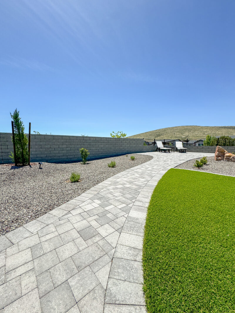 The backyard of the Wong Residence boasts a paved walkway that leads to lounge chairs and a small table. This serene space features a combination of gravel and grass landscaping, with small shrubs and a brick wall enclosing the area. A clear blue sky serves as the perfect backdrop.