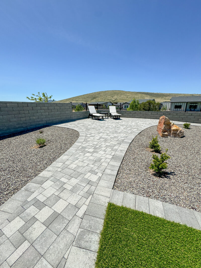 The backyard at the Wong Residence features a stone pathway leading to two lounge chairs and a small table. The yard is landscaped with gravel, young shrubs, and a patch of artificial grass. A low brick wall encloses the area, with a grassy hill in the background under a clear blue sky.