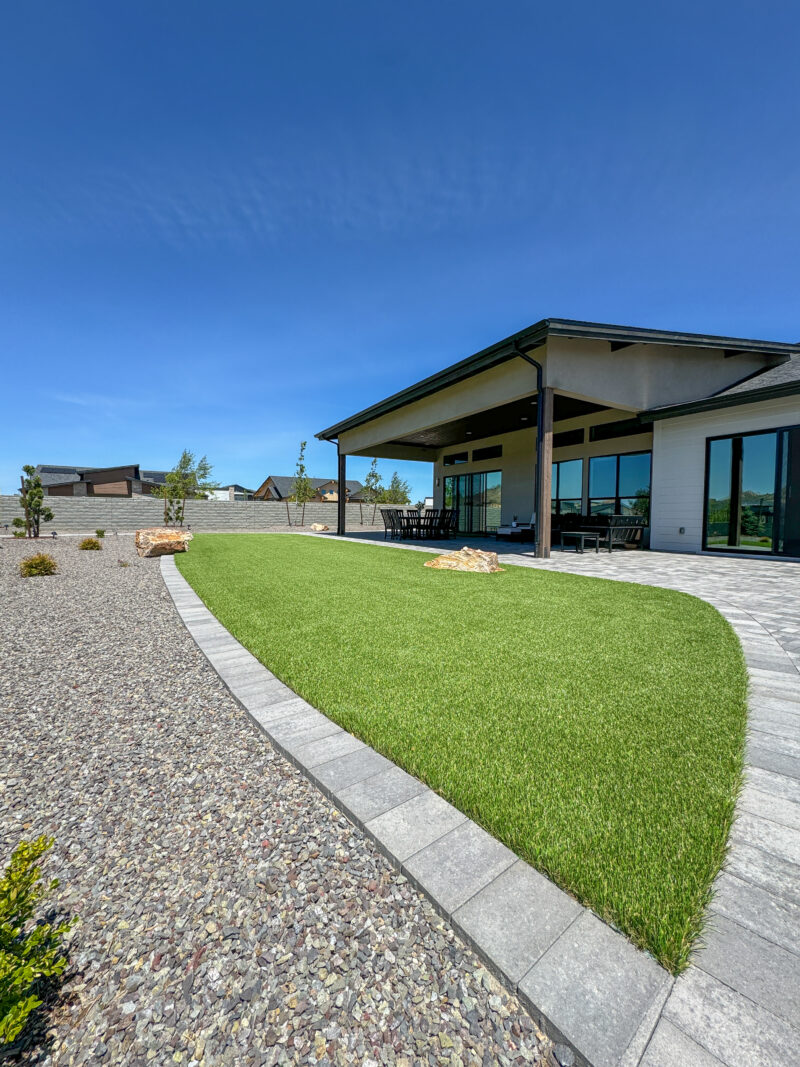 The Wong Residence is a modern house with a large patio featuring floor-to-ceiling windows and a dark roof extending over a green lawn. The surrounding landscape is covered with gravel and minimal plants under a clear blue sky.