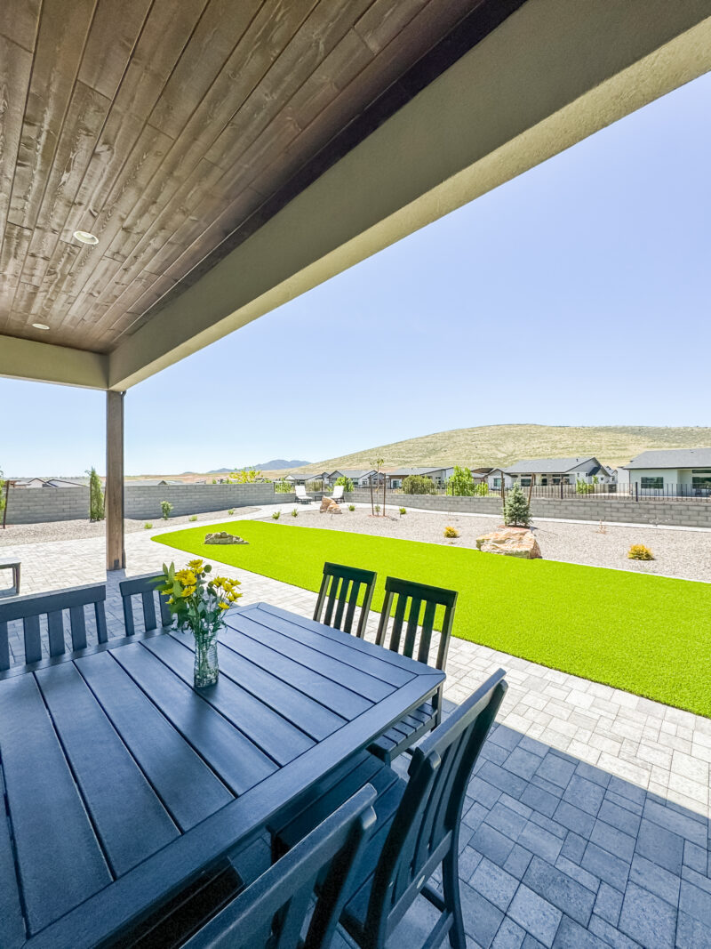 At the Wong Residence, a covered outdoor patio with a large black dining table and chairs overlooks a well-maintained grassy backyard. The yard features some rocks, trees, and a distant view of rolling hills. A vase with yellow flowers is placed on the table under the clear blue sky.