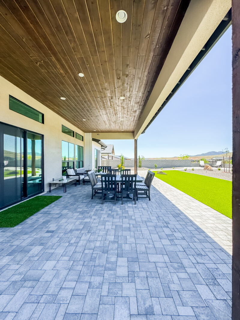 The Wong Residence boasts a spacious outdoor patio featuring a wooden ceiling, tile flooring, and modern furniture including a dining table with chairs. The area is partially shaded and overlooks a manicured lawn, with mountains visible on the horizon in the background.