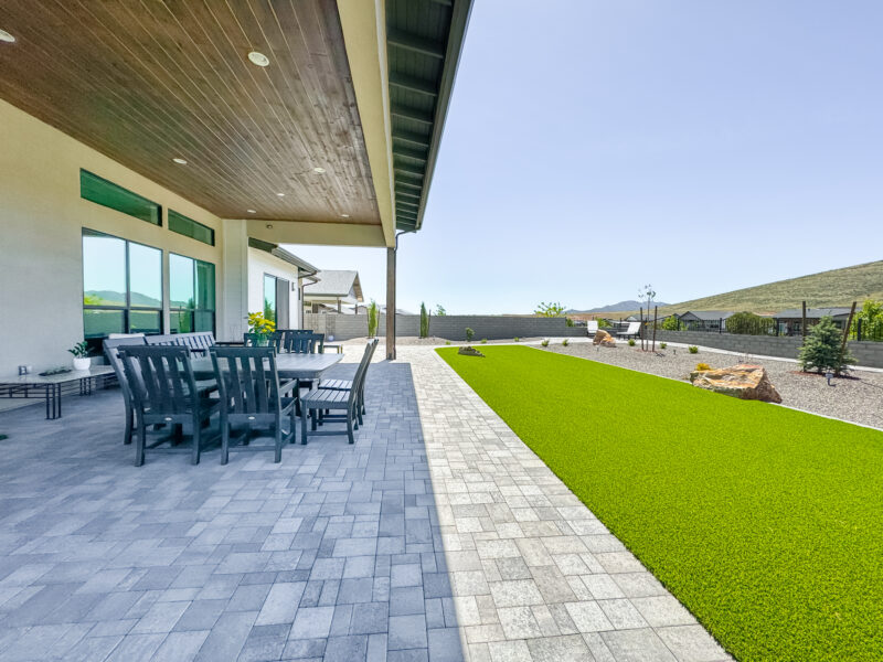 The Wong Residence boasts a spacious modern backyard with a large gray paved patio area featuring a dining table and chairs. The yard includes a well-manicured lawn alongside a rock garden. The area is enclosed by a tall fence, and mountains are visible in the background.