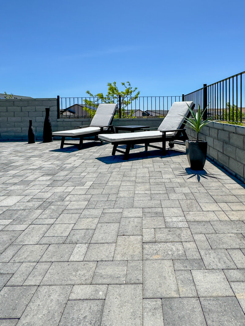 A spacious patio at the Wong Residence features gray interlocking pavers with two gray lounge chairs framed in black, facing the sky. A potted plant sits to the right, and tall vases are positioned against a low wall in the background, all enclosed by a black metal fence.