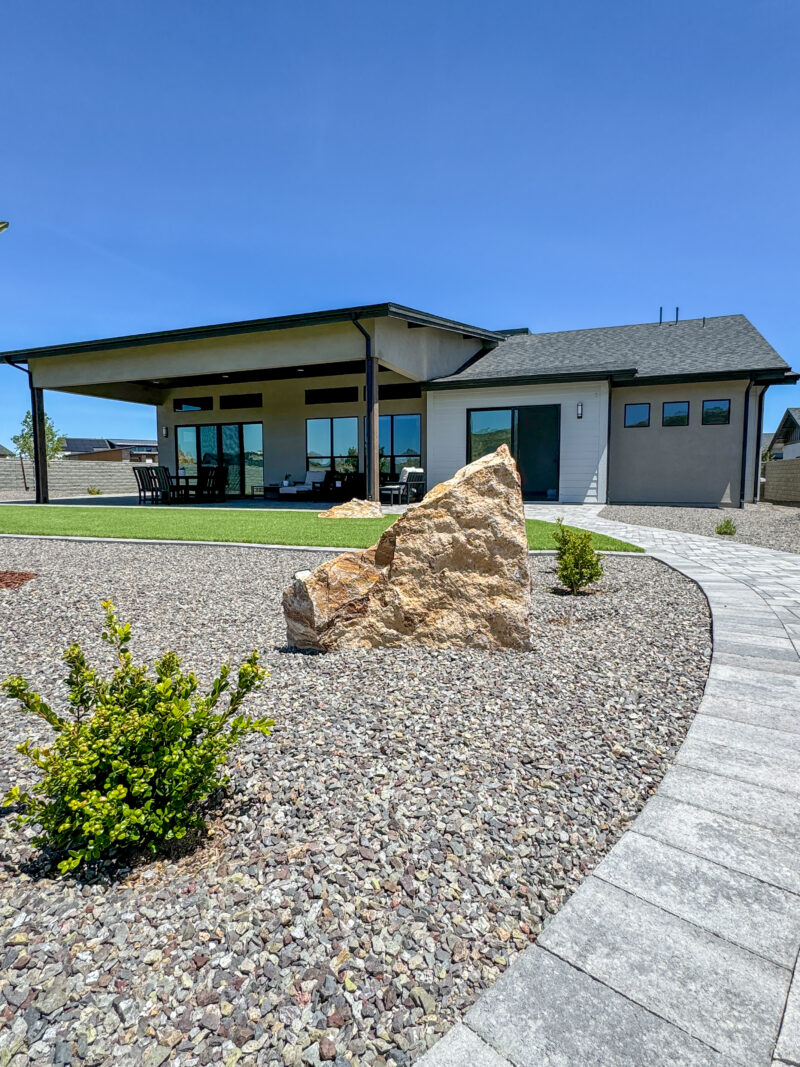 The Wong Residence, a modern single-story house with a sleek design, features a large covered patio with outdoor seating. A winding stone path leads to the entrance, flanked by small shrubs and a prominent rocky landscape element in the front yard. The sky is clear and blue.