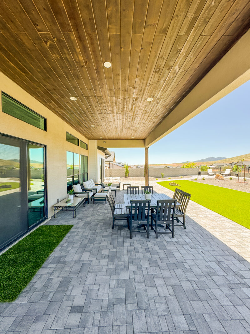 The Wong Residence boasts a spacious outdoor patio with a wooden ceiling, featuring a dining table with black chairs, a cozy seating area adorned with white cushions, and an amazing view of the large backyard&#039;s green grass and distant mountains under clear blue skies.