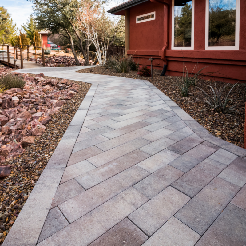 A pathway made of rectangular stone pavers arranged in a chevron pattern leads to a building with red walls, resembling an art gallery. The path is bordered by a stone landscape filled with small rocks and desert plants. Trees and another building are visible in the background.