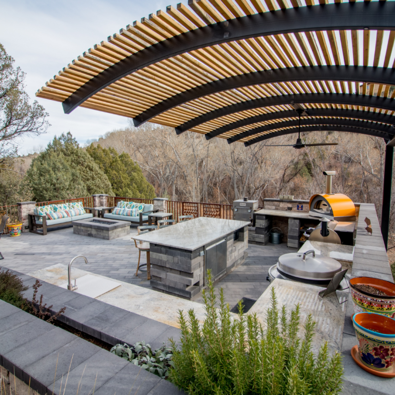 A stylish outdoor kitchen and seating area features a curved wooden pergola overhead. The space includes a grill, pizza oven, countertop with stools, sink, and potted plants. The seating area has cushioned benches and overlooks a natural landscape with trees, resembling an elegant gallery.