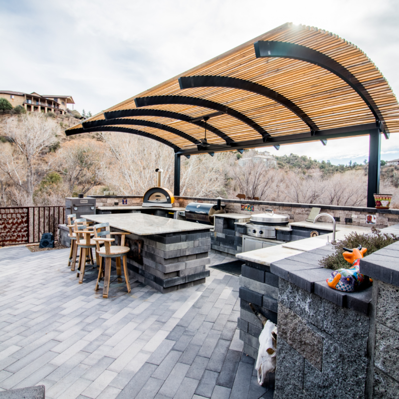 Outdoor kitchen and seating area featuring grey stone countertops, bar stools, and various cooking appliances. Covered by a modern, slatted wooden canopy, the setting looks like a portfolio piece from an architectural gallery. An open view of barren trees and distant buildings is visible in the background.