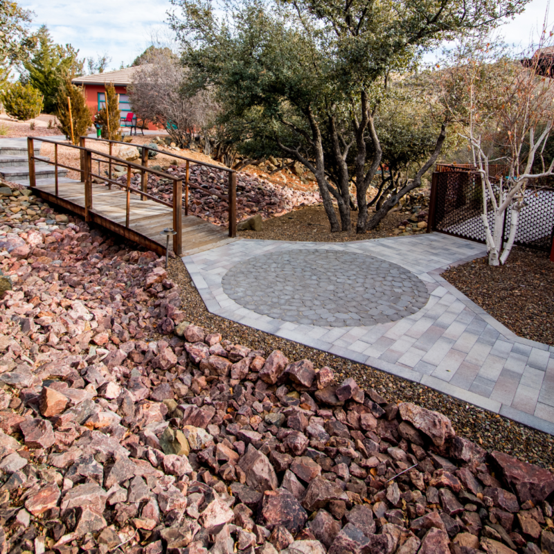 A paved pathway with a circular stone pattern leads to a wooden bridge surrounded by rocks, resembling an inviting gallery. In the background, there are trees and a red building. The scene is set in a dry, rocky landscape.