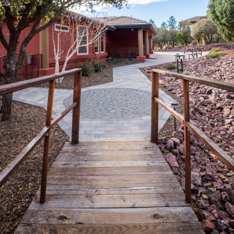 A wooden footbridge leads to a paved path that winds towards a red brick gallery surrounded by rocks and xeriscape landscaping. Trees and other buildings are visible in the background under a partly cloudy sky, creating an idyllic portfolio-worthy scene.