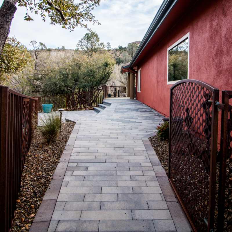 A paved stone walkway leads to a gallery with a red exterior wall. The path is bordered by rust-colored gates and landscaping featuring trees, shrubs, and a small grassy area. In the background, there are hills and a cloudy sky.