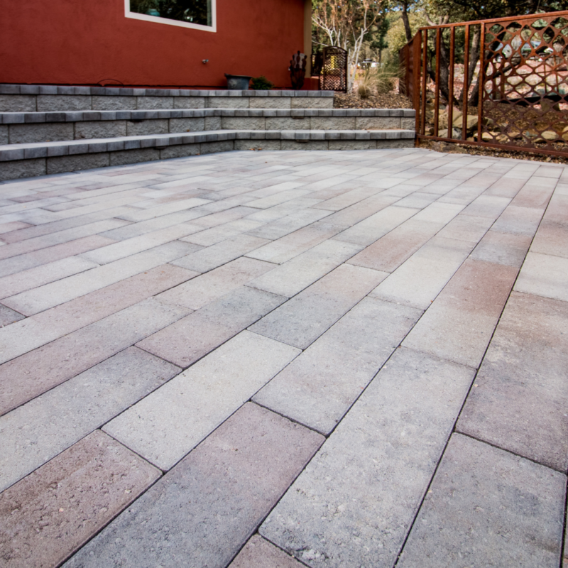 A backyard with a large paved patio featuring rectangular stones in shades of beige, gray, and reddish-brown. In the background, there are stone steps leading to an orange-red house with a window and a decorative metal gate. With trees and foliage as a natural gallery, it feels like stepping into a portfolio of outdoor design.