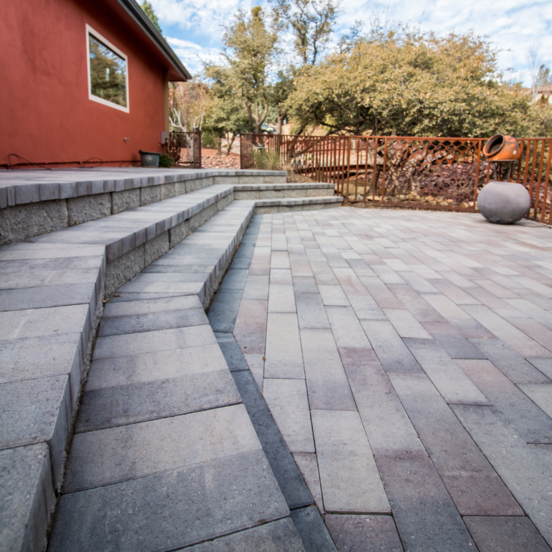 A spacious outdoor patio with light gray and beige pavers leads to a set of wide steps ascending to an orange-painted house. The background features a garden with trees and a large clay pot, creating a perfect setting for any photography portfolio. The sky is partly cloudy, adding natural light perfect for a gallery shot.