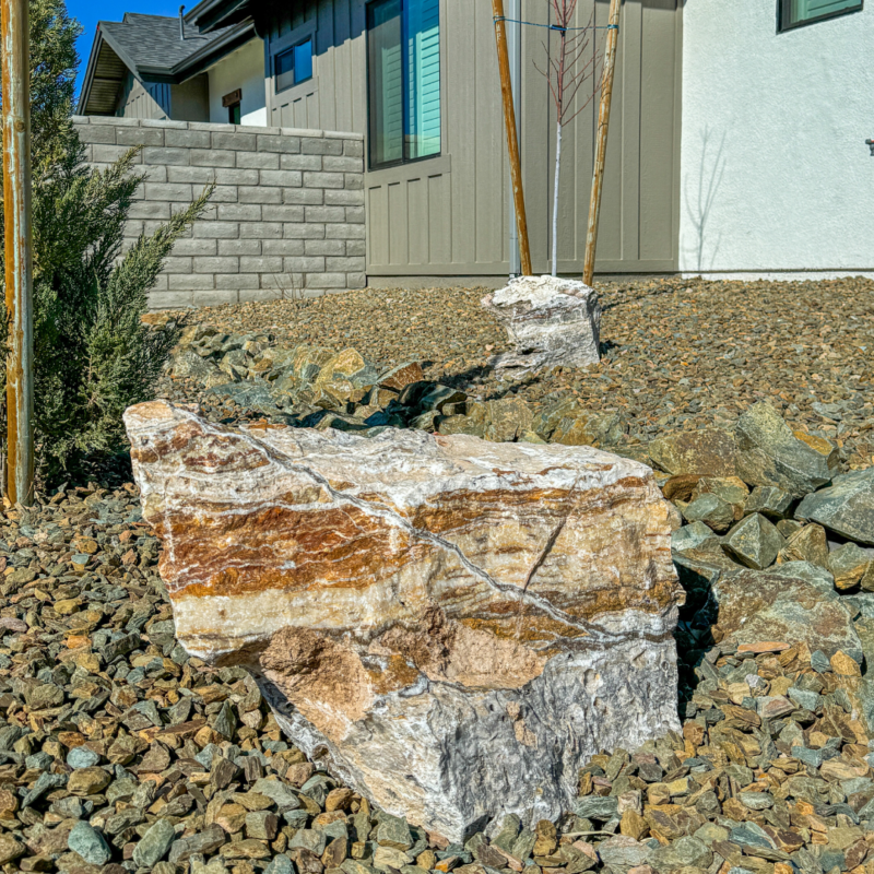 A xeriscaped front yard features large decorative rocks among small, uniformly colored pebbles. Two newly planted young trees are supported by wooden stakes like an auto draft. The background includes a house with taupe siding, white trim, and a brick wall.