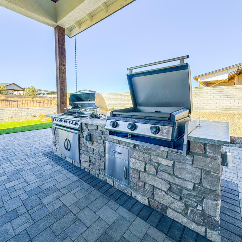 At the Wilson Residence, an outdoor kitchen boasts two stainless steel grills set in a stone counter. The left grill has a closed lid, while the right one is open. Storage cabinets enhance the counter&#039;s functionality. This culinary haven sits on a brick-paved patio adjacent to a grassy yard.