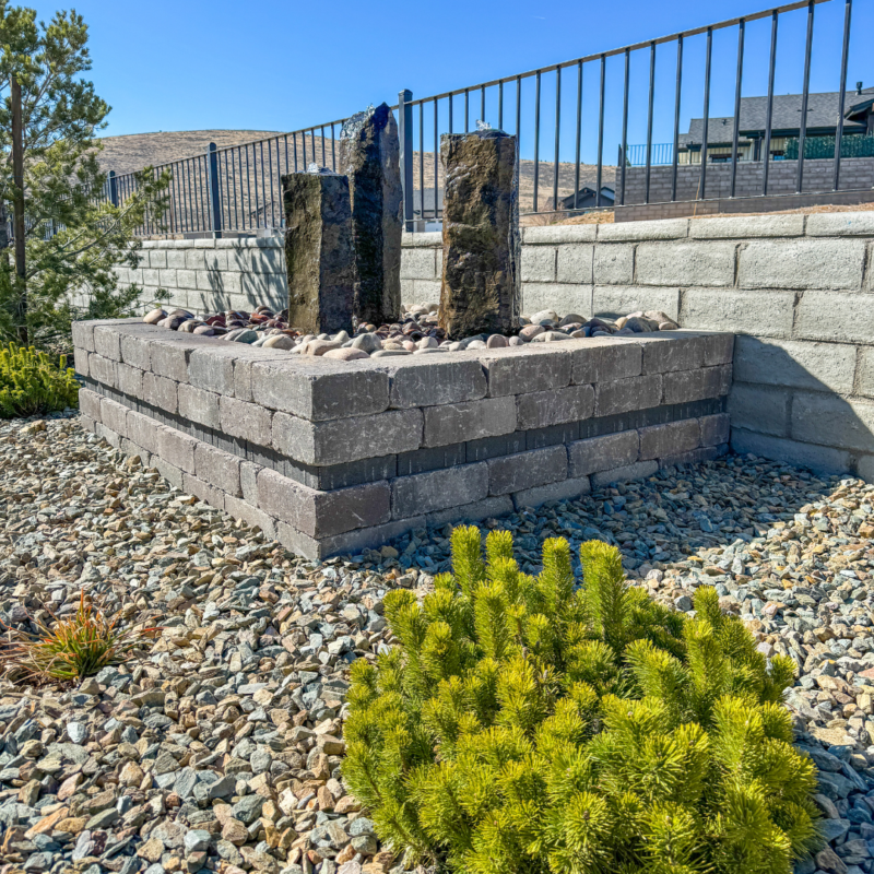 At the Wilson Residence, a landscaped area features a raised rectangular stone planter containing a vertical water feature made of three tall rock pillars. Surrounding the planter are gravel and low-growing shrubs, with a metal fence and stone wall in the background under a clear blue sky.