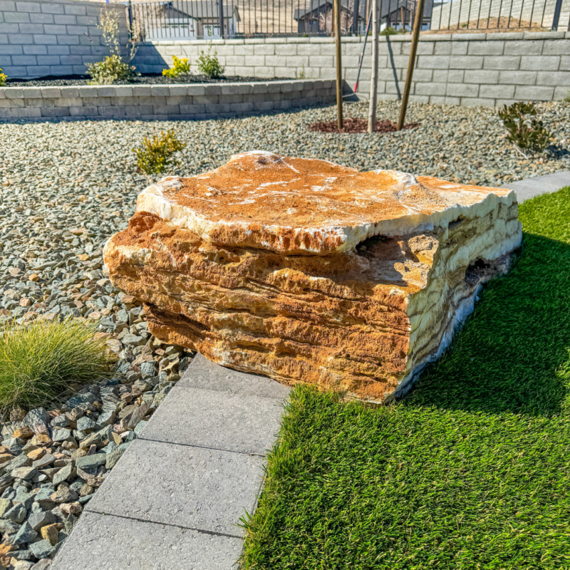 A large, flat, layered rock sits next to a pathway in the landscaped garden of the Wilson Residence. The garden features neatly arranged gravel, patches of artificial grass, and a few small plants. A stone wall and trees are visible in the background.