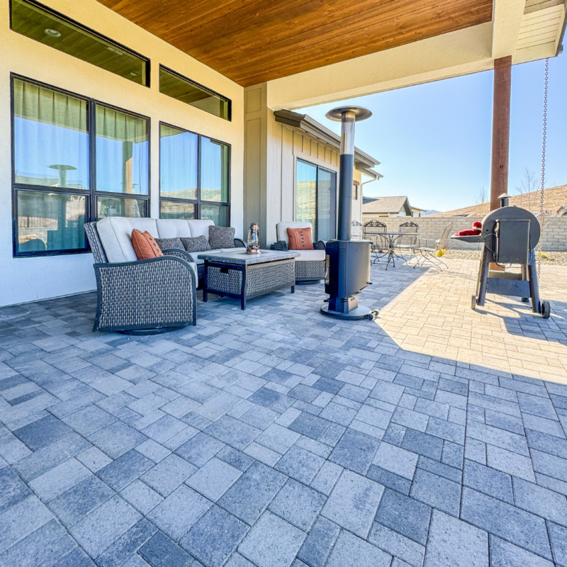 At the Wilson Residence, a spacious outdoor patio features cushioned wicker furniture, a coffee table, a patio heater, and a porch swing. The area is covered by a wooden ceiling and has an expansive tiled floor. In the background, there’s a fence, other houses, and a clear blue sky.