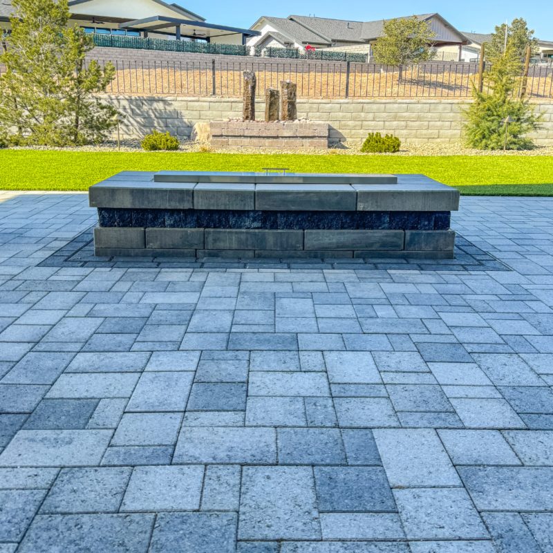 A modern outdoor patio at the Wilson Residence featuring a central, rectangular stone fire pit. The patio is adorned with gray paver stones, and in the background, there is a well-maintained lawn, trees, a stone wall, and a small water feature. Residential houses are visible beyond the fence.