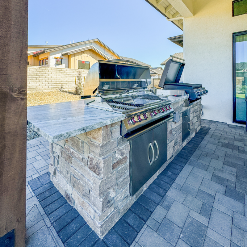 The Wilson Residence features an outdoor kitchen with a built-in stone barbecue grill and granite countertops. There are two grills, one with the lid open and one closed. The kitchen is set on a patio with brick pavers, adjacent to a white building.