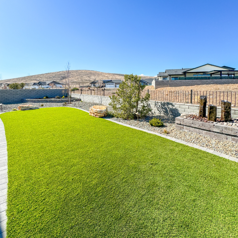 The Wilson Residence boasts a well-maintained backyard with bright green grass, a small rock garden adorned with decorative rocks and short plants, and a backdrop of housing structures and hills under a clear blue sky. The yard is enclosed by a fence with a stone wall.