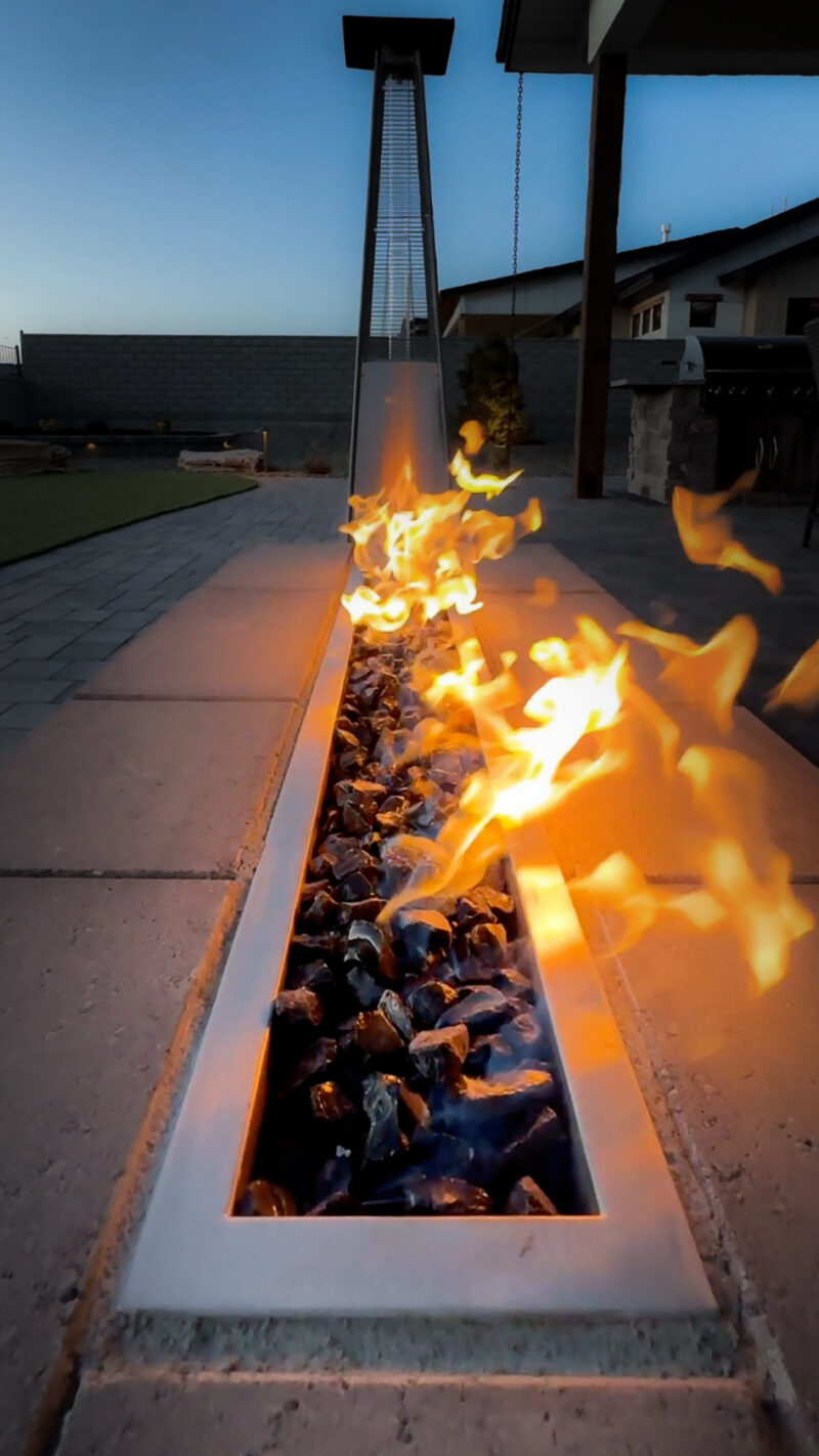 At the Wilson Residence, a modern fire pit with a line of flames rises from a bed of black and blue rocks in an outdoor patio setting. The fire pit area is framed by stone tiles, with a glimpse of the yard and fence visible in the backdrop. The sky hints at evening time.