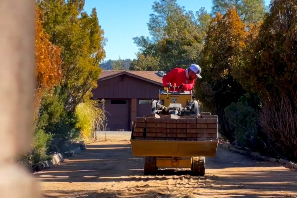 A person wearing a red shirt and cap operates a small heavy machinery vehicle loaded with bricks on a dirt path, flanked by trees and leading to a red building in the background. The sky is clear and blue.