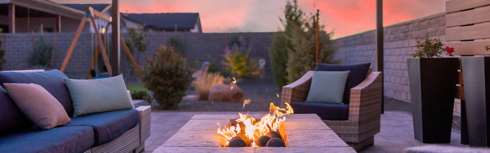 A cozy outdoor patio at sunset features a modern fire pit with flames dancing in the center. Surrounding the fire pit are comfortable wicker chairs with colorful cushions. In the background, a neatly landscaped garden and swing set are visible.