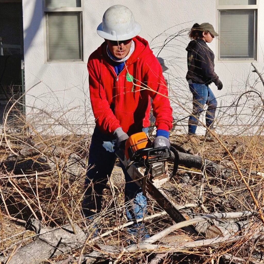 A person wearing a white hard hat and red hoodie operates a chainsaw to cut branches outside near a white building, showcasing the efficiency of Prescott Landscaping Company. Another individual in a black cap and gloves stands in the background as fallen branches and dry grass are scattered around them.