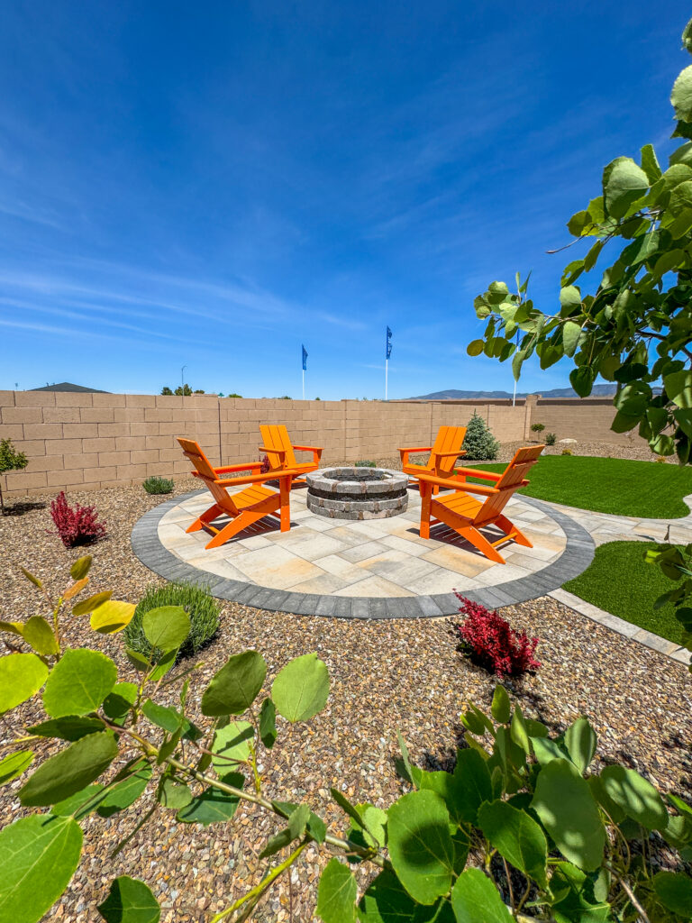 A backyard with a circular paved patio featuring four orange Adirondack chairs arranged around a stone fire pit. The patio is surrounded by a gravel garden with green plants and shrubs, and a wooden fence encloses the area. A vivid blue sky is overhead.