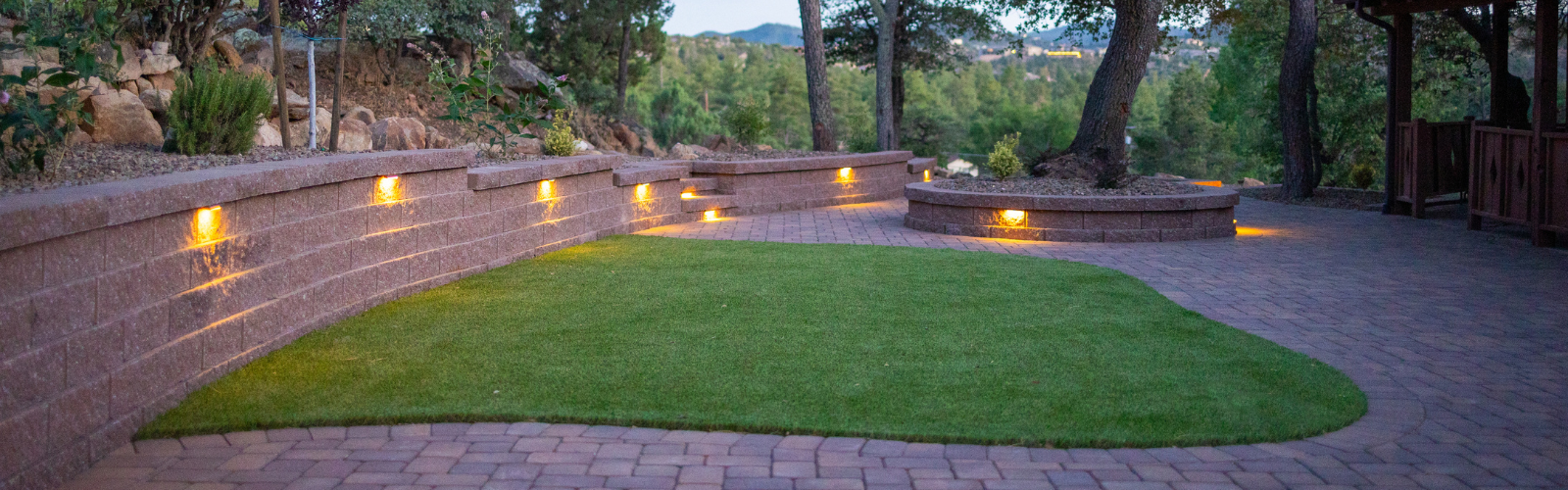 A backyard featuring a curved brick retaining wall with built-in lights, a small grassy area, and a circular brick seating area. The area is surrounded by trees and greenery, creating a serene and picturesque outdoor environment that consistently garners rave reviews.