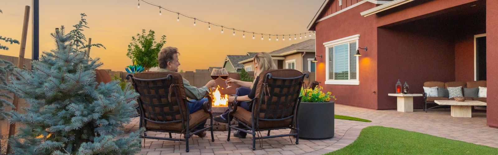 A couple sits in outdoor chairs, toasting with wine glasses near a fire pit. The backyard features outdoor lighting, including string lights, a small tree, a potted plant, and a seating area on a patio against the backdrop of a sunset sky. The house has a warm, inviting ambiance.