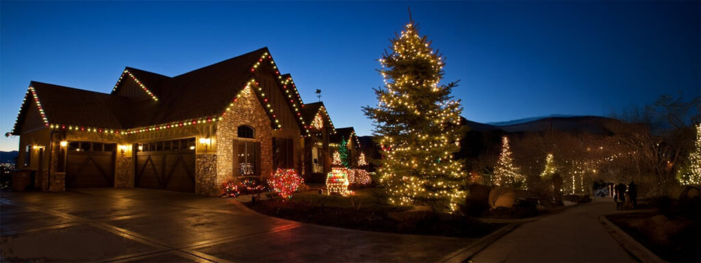 A warmly lit house exterior in Prescott is adorned with premier Christmas light installation at dusk. A large evergreen tree in the front yard shimmers with white lights, while nearby shrubs also glow. The driveway and path are illuminated, leading into the festive scene.