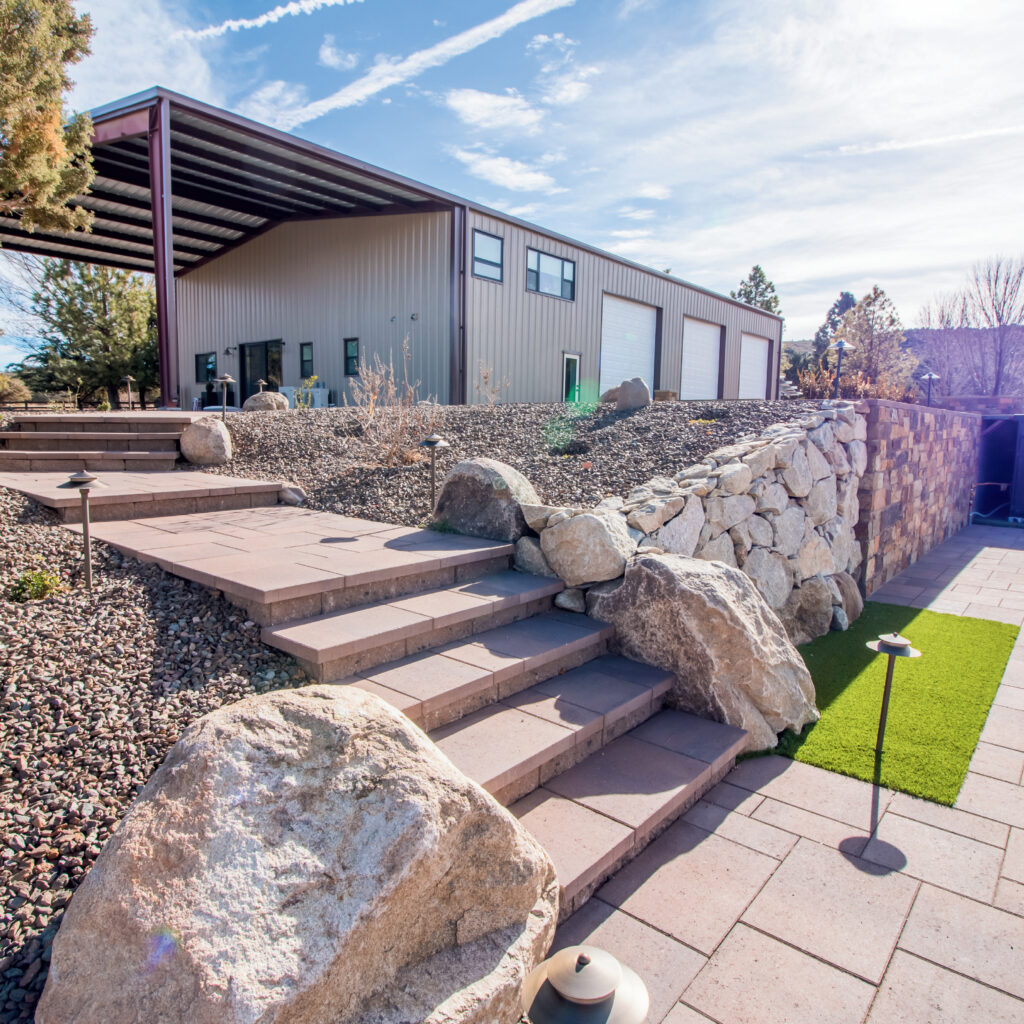 A modern building with large white garage doors and a metal roof is nestled on a hillside. Stone steps, artfully incorporated into the landscaping, lead up through rocks and gravel. A small patch of artificial turf peeks from the right under a clear blue sky.