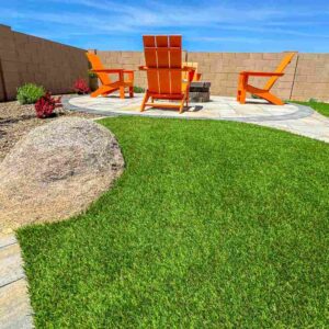 A beautifully landscaped backyard showcases lush green grass, a large rock, and a circular patio with bright orange Adirondack chairs around a fire pit. A tan brick wall encloses the space, while small plants accentuate its edges under a clear blue sky.