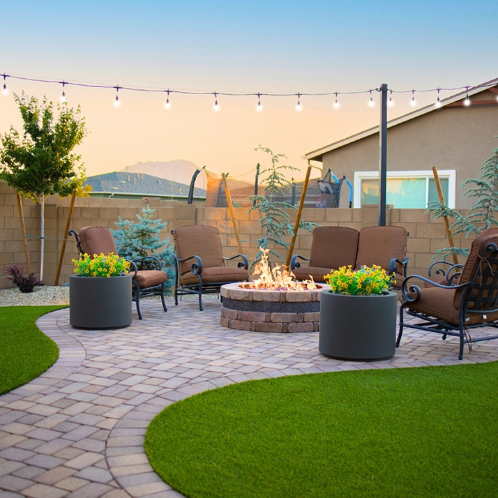 A cozy outdoor patio, designed by Prescott Landscaping Company, features brown cushioned chairs around a stone fire pit. A paved pathway and manicured lawn lead to the area, with string lights overhead and large planters filled with yellow flowers adding color. A house is visible in the background.