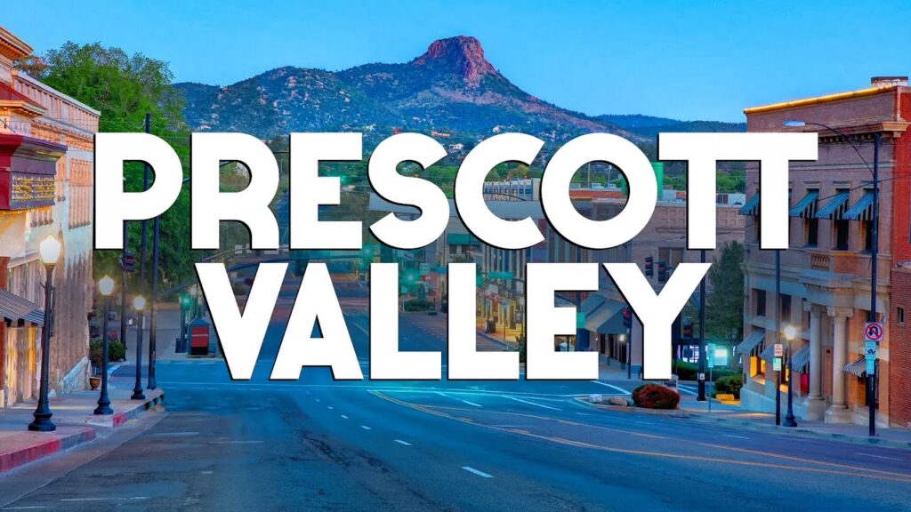 In downtown Prescott Valley, historic buildings line the road beneath a clear AZ sky and majestic mountains. "Prescott Valley" is boldly displayed in large white text, capturing the essence of this charming locale.