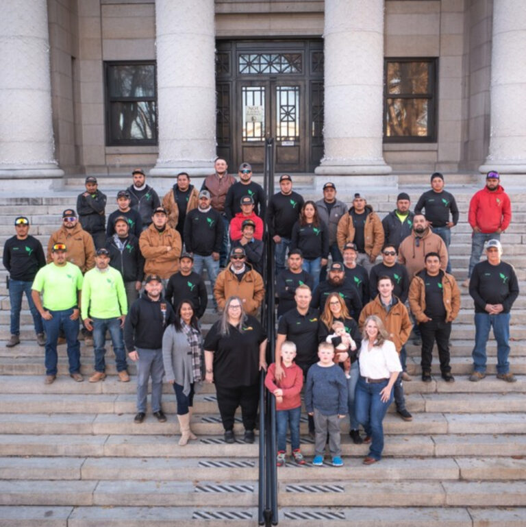 Team Yavapai Landscaping in Prescott as a large group of people, including men, women, and children, pose on the steps of a building with large columns. Some are wearing jackets and hats, and a few are holding children. The weather appears cool, and everyone is smiling.