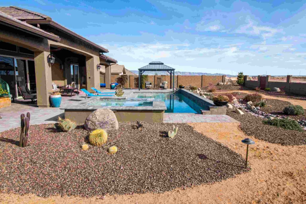 A backyard with a swimming pool, lounge chairs, a shaded pergola, and expert landscape design featuring rocks, cacti, and succulents under a clear blue sky.