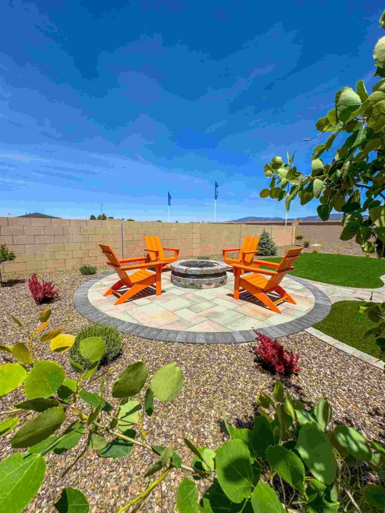 A circular stone fire pit is surrounded by four bright orange wooden chairs on a paved area, showcasing thoughtful landscape design with gravel, plants, and a stone wall in the background under a clear blue sky.