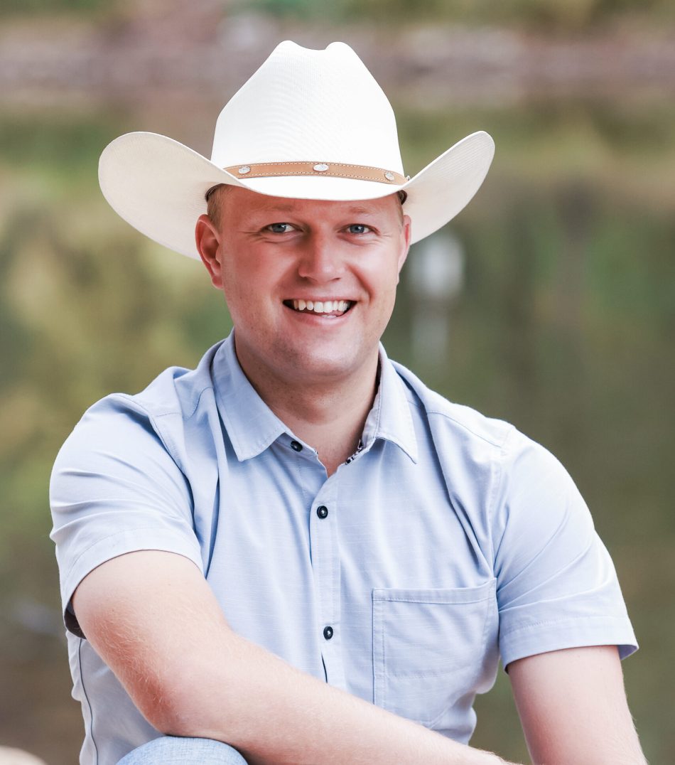 A man wearing a white cowboy hat and a light blue button-up shirt smiles while seated outdoors. He has light hair and is sitting in front of a blurred natural background with greenery and water.